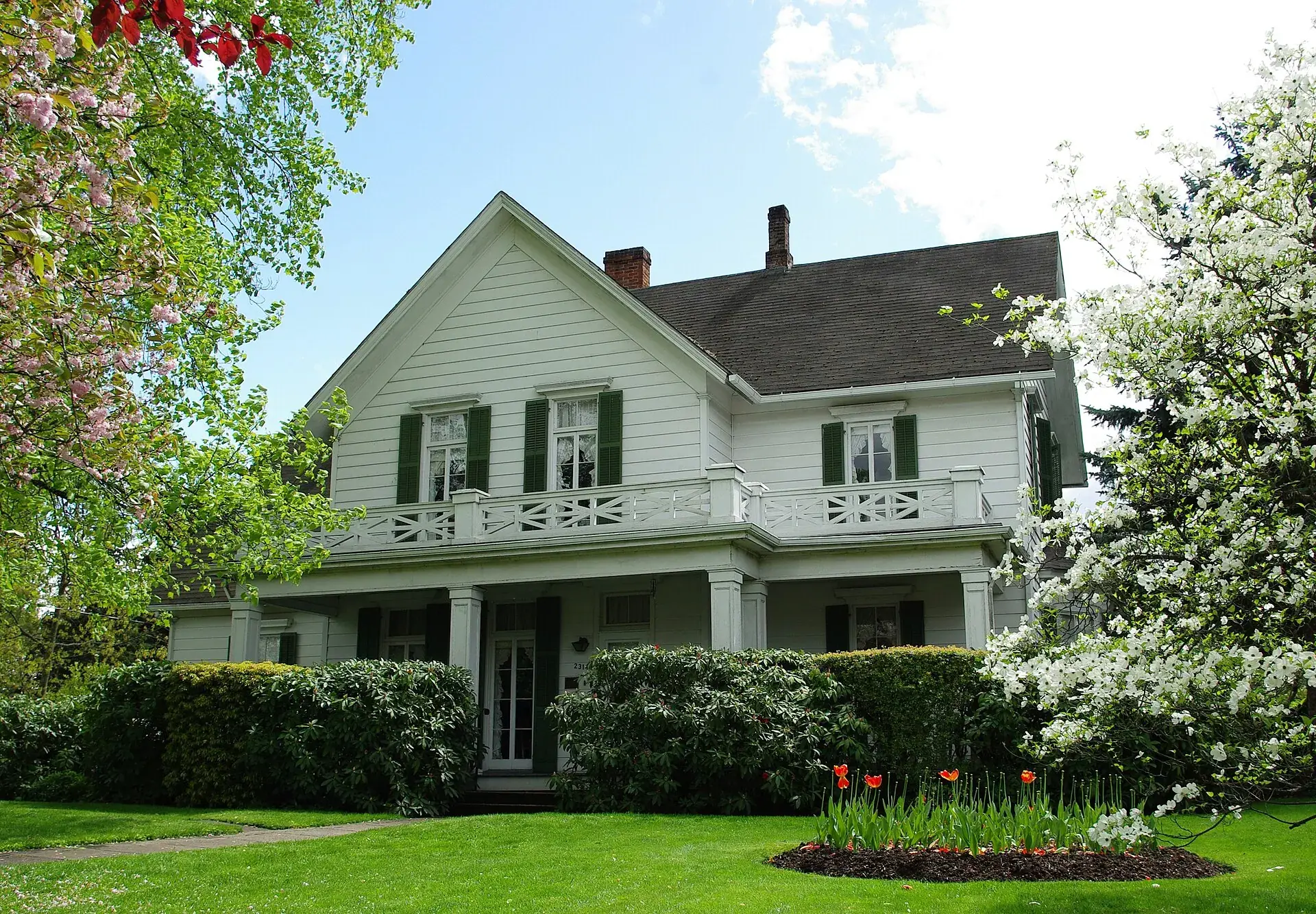 A white 19th-century Oregon farmhouse with green shutters and a covered porch, framed by flowering trees